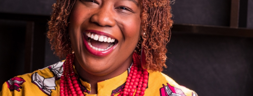 Head shot of Dr. Crystal Moten with gold top, red bedded necklace, smiling as if laughing.