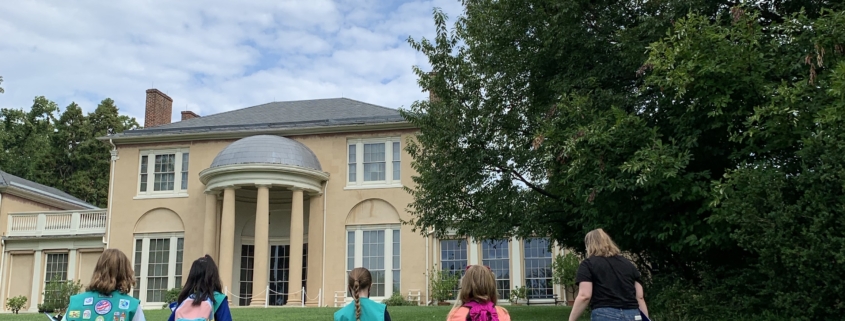 Girl Scouts walk up the South Lawn towards the historic house with their green Girl Scout sashes on.